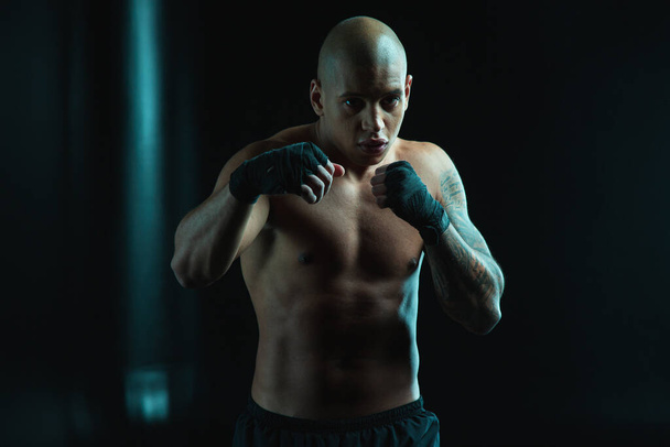 Muscular tattooed bold man looking at you while standing in boxing pose with bandages on the fists - Fotografie, Obrázek