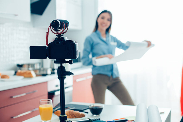 Confident blogger standing in the kitchen with roll of architectural paper and recording a video with a help of a camera on her table - Foto, Imagem
