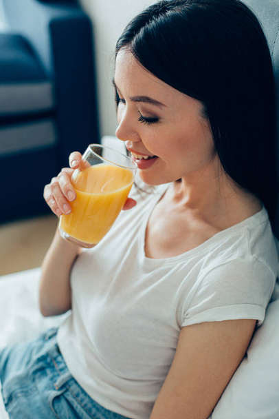 Cheerful beautiful young lady sitting at home and looking happy while drinking orange juice from the glass - Fotoğraf, Görsel