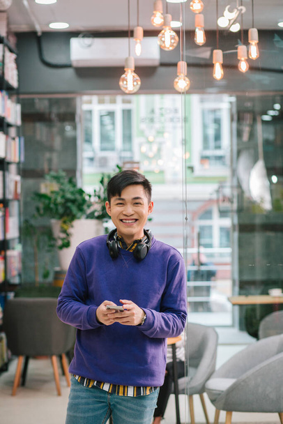 Cheerful young Asian man in casual clothes standing in the library with bright illumination behind him and smiling while holding a smartphone - Photo, image