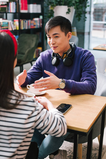 Cheerful young man with headphones on the neck sitting at the cafe table and gesturing while talking to a young lady - 写真・画像