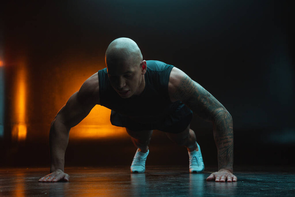 Concentrated attractive athlete with tattooed arm looking serious while doing the push ups. Orange light on the background - Фото, зображення