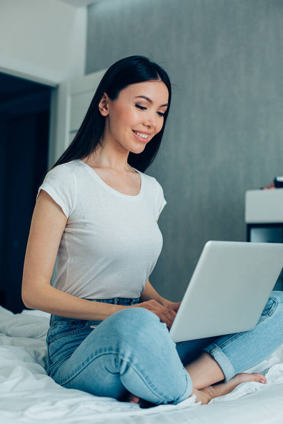 Attractive young Caucasian woman sitting on a bed in jeans and white T-shirt and using a modern laptop - Foto, Bild