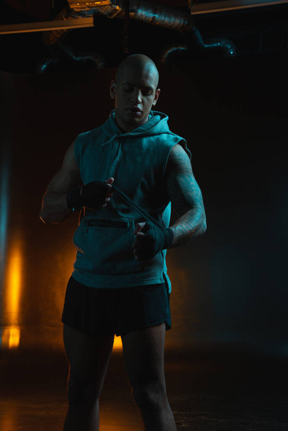 Calm muscular man looking concentrated while standing in the dark studio and tying boxing bandages - Foto, Bild