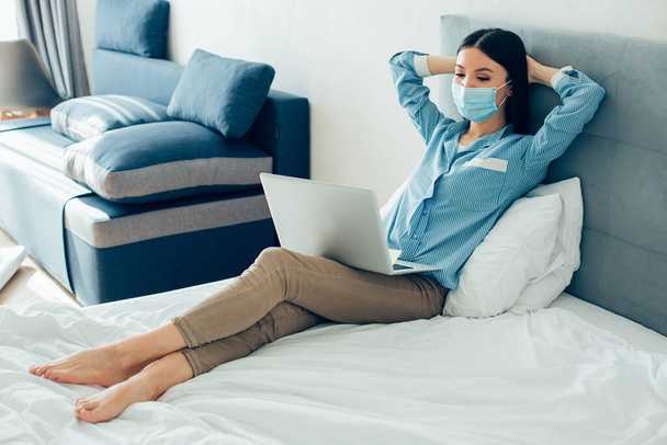Молодая босоногая женщина в медицинской маске сидит на кровати с руками за головой и смотрит на экран ноутбука - Фото, изображение