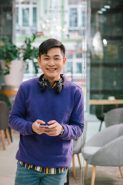 Contented young Asian man standing alone with a modern smartphone in his hands and smiling. Room with glass door behind his back - Foto, Bild