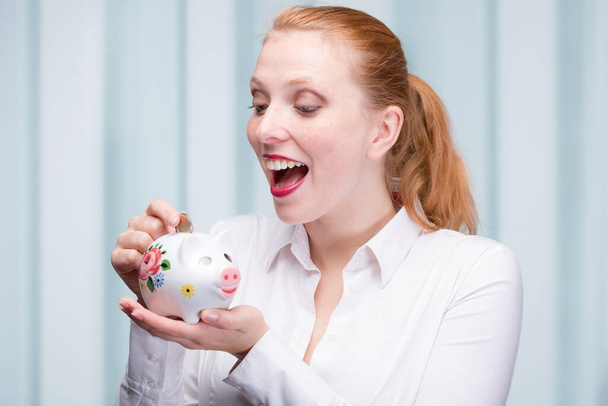 young red haired woman puts a coin into her piggy bank / porcela - Foto, Imagen