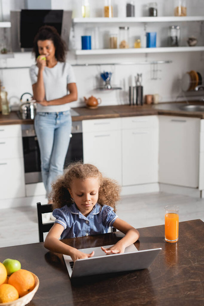 concentrated african american girl using laptop while sitting at kitchen table near mother standing on background - 写真・画像