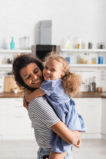 african american girl looking at camera and embracing mother holding her on hands in kitchen - Zdjęcie, obraz