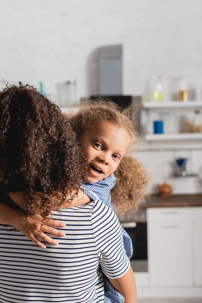 Вид сзади женщины в полосатой футболке, держащей в руках африканскую дочь, пока она обнимает ее и смотрит в камеру - Фото, изображение