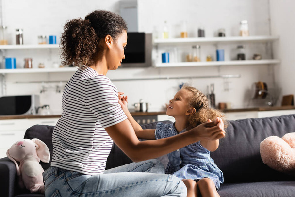 african american woman in striped t-shirt holding hands with daughter while having fun on sofa in kitchen - Фото, зображення