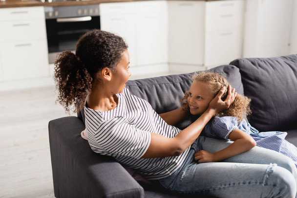 young african american mother touching head of excited daughter while sitting on sofa at home - Foto, afbeelding