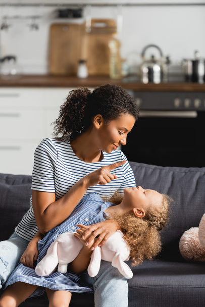 fiatal afro-amerikai anya csíkos pólóban szórakozik a lányával, miközben a kanapén ül a konyhában - Fotó, kép