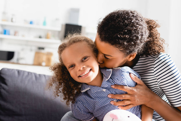young african american mother kissing neck of excited daughter looking at camera - Foto, afbeelding