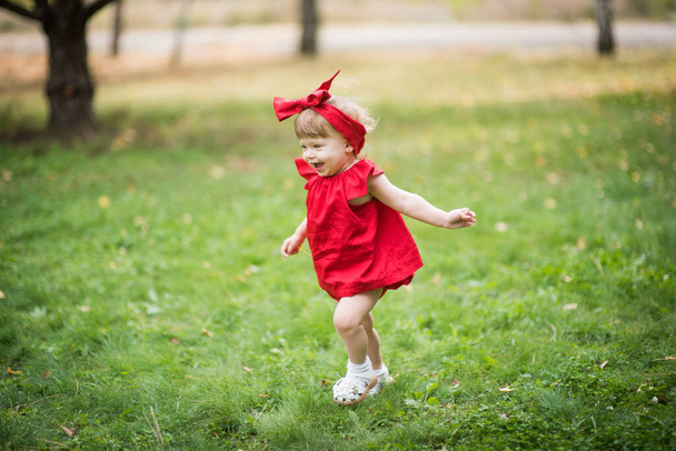 pequeña chica rubia en vestido rojo sonrisa y correr sobre hierba verde - Foto, imagen