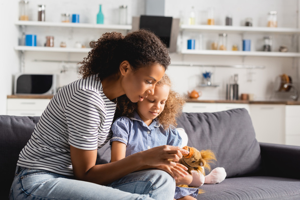 young african american babysitter touching toy in hands of girl sitting on couch in kitchen - Photo, Image