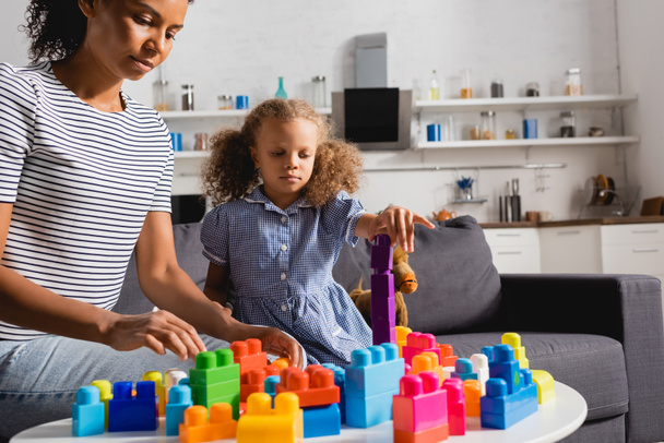 afro-américaine fille en robe et baby-sitter en t-shirt rayé jouer avec des blocs de construction colorés dans la cuisine - Photo, image