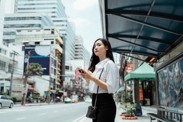 Asijské podnikatelka na bílý shirt hrát smartphone a čeká na autobus na autobusové zastávce. - Fotografie, Obrázek