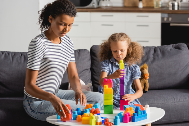 young african american babysitter and girl playing with colorful building blocks while sitting on sofa - Photo, Image