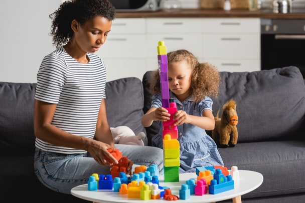 african american kid building tower from multicolored blocks while sitting on couch near nanny - Photo, image