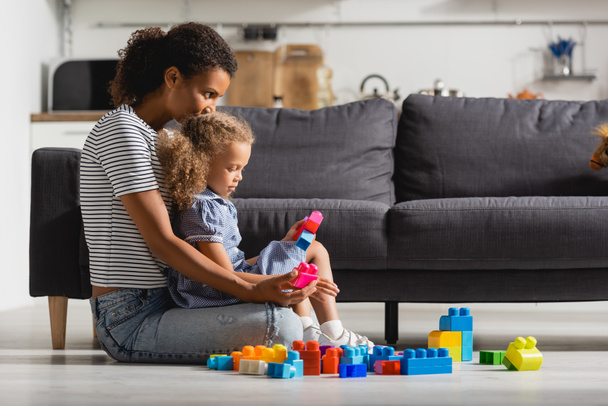 side view of african american girl playing with multicolored building blocks on floor near nanny - 写真・画像