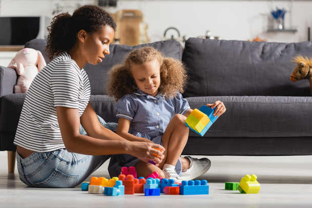 african american girl and nanny playing with colorful building blocks on floor near sofa - Photo, Image