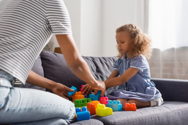 partial view of babysitter and african american girl playing with colorful building blocks on couch - Photo, Image