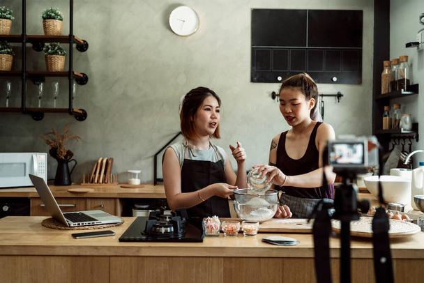 Zwei junge asiatische Bäckerinnen backen Brot in der Küche. Aufzeichnung von Videos mittels Kamera im Stativ für eine Kochshow. Vlogging-Video über Brot backen. - Foto, Bild