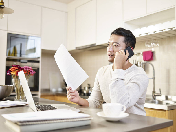 junger asiatischer Geschäftsmann sitzt am Küchentisch und arbeitet zu Hause und telefoniert mit einem Dokument - Foto, Bild