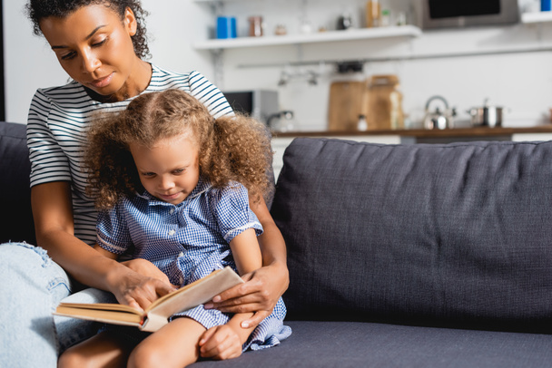young african american nanny and concentrated child sitting on couch and reading book together - Фото, изображение