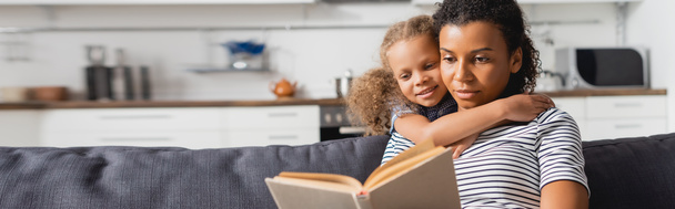 Afro-Amerikan bir kızın yatay görüntüsü. Kanepede oturmuş mutfakta kitap okuyor. - Fotoğraf, Görsel