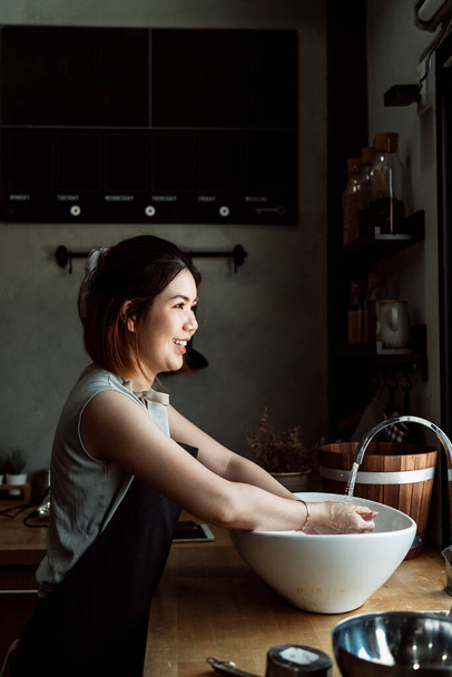 Весела азіатська жінка миє руки біля водяної мийки перед приготуванням на кухні
. - Фото, зображення