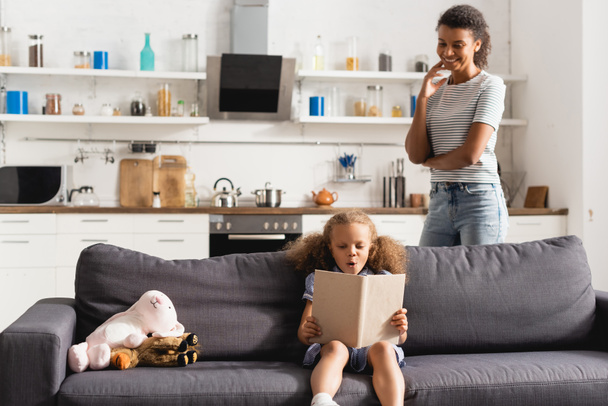 joven afroamericano niñera de pie detrás excitado chica lectura libro mientras sentado en sofá cerca de juguetes - Foto, Imagen