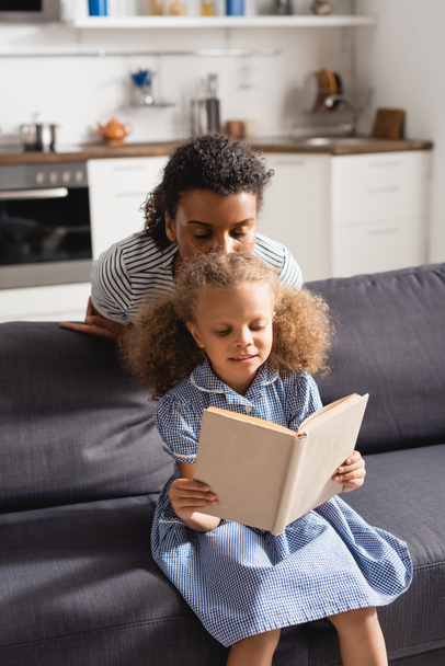 african american woman with closed eyes kissing head of daughter reading book on sofa - Photo, Image
