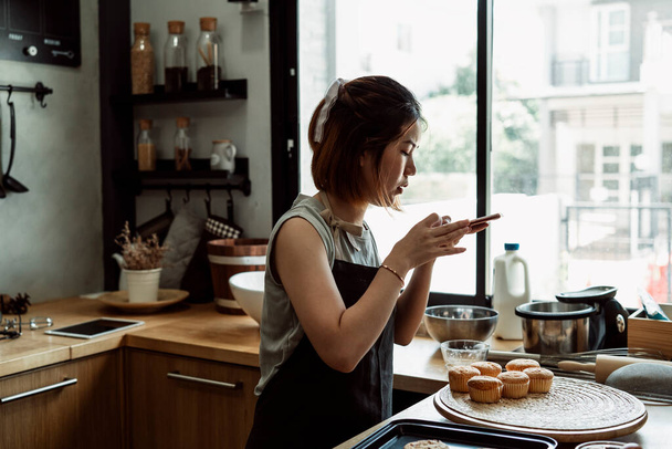 Panadera asiática tomando fotos de magdalenas frescas en un plato de madera en la cocina con smartphone para redes sociales. - Foto, Imagen