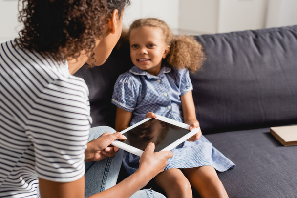 selective focus of babysitter giving digital tablet with blank screen to african american girl sitting on couch  - Foto, Imagen