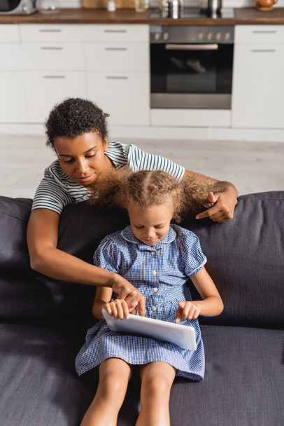 vista de ángulo alto de la niñera afroamericana apuntando con el dedo a la tableta digital en manos de un niño sentado en el sofá - Foto, Imagen