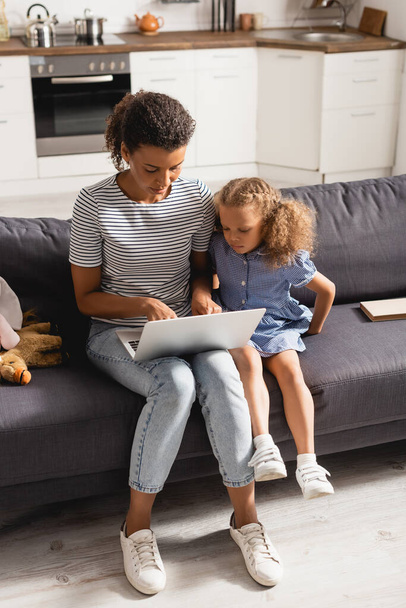 high angle view of african american freelancer in striped t-shirt and jeans using laptop near daughter on sofa - Foto, Imagem