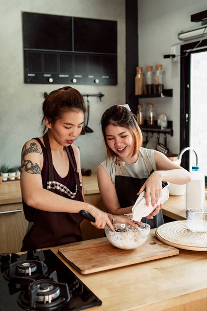 Két ázsiai péknő lisztet, tejet, tojássárgáját keveri üvegtálba a konyhában. Kenyérkészítési eljárás. - Fotó, kép