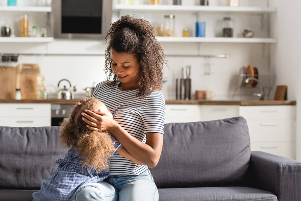 若いアフリカ系アメリカ人の女性が家でソファに座っている間に娘の頭に触れる縞模様のTシャツ - 写真・画像