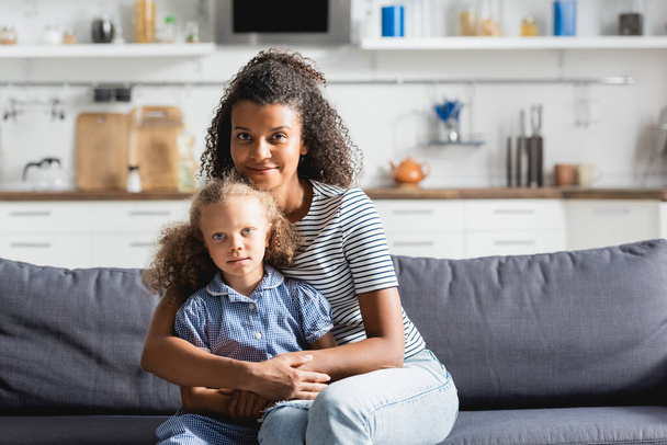 młoda afrykańska amerykanka obejmująca córkę i patrząca w kamerę siedząc na kanapie w kuchni - Zdjęcie, obraz