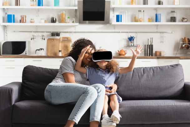 young african american nanny embracing child in vr headset gesturing on sofa in kitchen - Foto, afbeelding