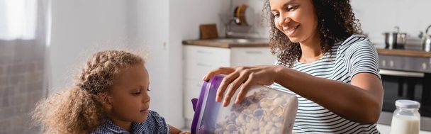 horizontales Bild einer jungen afrikanisch-amerikanischen Frau, die Container mit Cornflakes in der Nähe ihrer Tochter hält - Foto, Bild