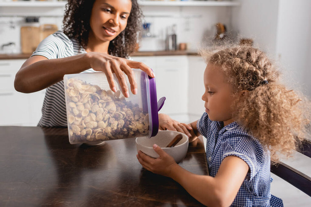 selective focus of african american woman pouring cornflakes from container into bowl near daughter - Fotoğraf, Görsel
