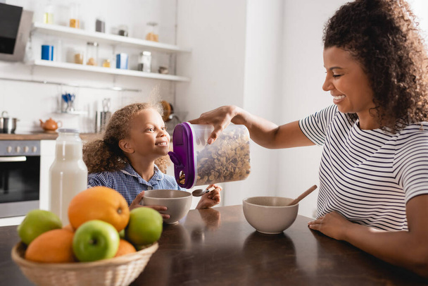 selective focus of young african american woman in striped t-shirt pouring cornflakes into bowl near excited daughter - Foto, Imagen