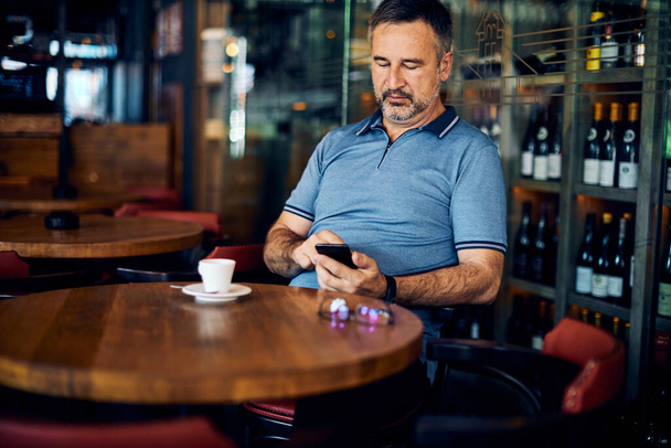 Mature man sitting in cafe and using smart phone. There is a coffee on a table. - Photo, Image