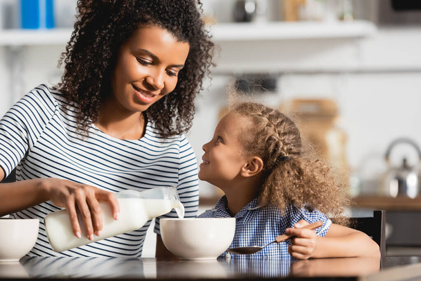 young african american woman in striped t-shirt pouring fresh milk into bowl near daughter holding spoon - Foto, immagini