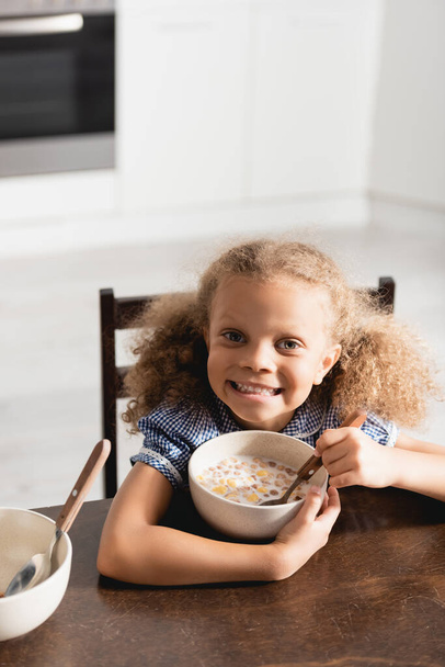 vista de ángulo alto de la chica afroamericana emocionada mirando a la cámara mientras sostiene la cuchara cerca del tazón con leche y copos de maíz - Foto, imagen