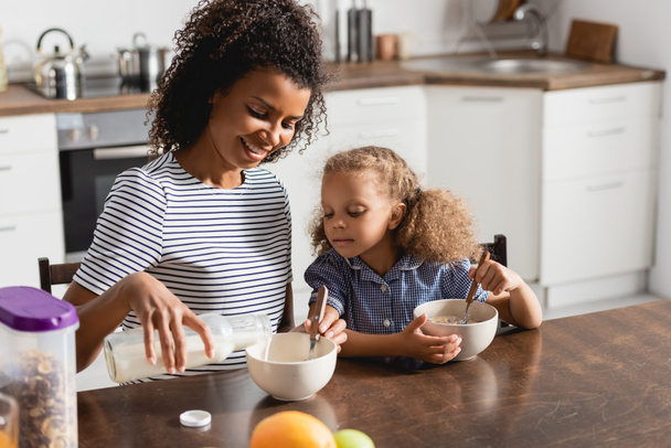 african american mom in striped t-shirt pouring milk into bowl while having breakfast with daughter - Foto, immagini