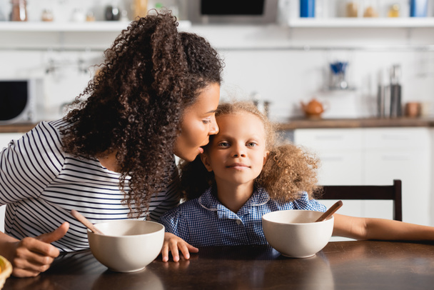 femme afro-américaine en t-shirt rayé chuchotant à l'oreille de sa fille pendant le petit déjeuner - Photo, image